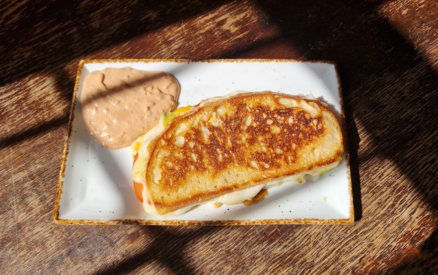 auf einer alten tischplatte steht ein weißer rechteckiger teller im sonnenschein der durch ein fenster scheint.
auf dem teller liegt ein gegrilltes dinkel sandwich man sieht das käse und tomaten aus dem sandwich ragt . das sandwich wurde goldbraun angebraten
dazu ist ein cocktaildip auf dem teller angerichtet