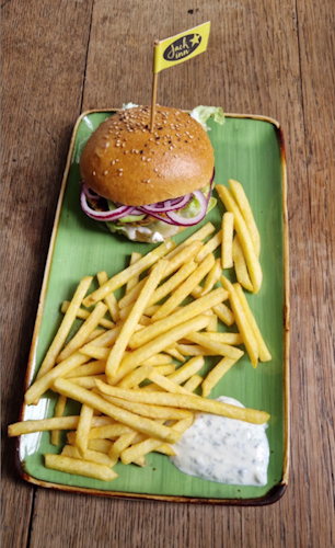 auf einem alten tisch steht ein länglich rechteckiger teller mit einer joiughurtsoße und pommes.
am oberen rand des tellers liegt ein falafelburger. man sieht ein paar rote zwiebeln und etwas gurke. das brötchen hat eine schöne braune farbe mit sesam bestreut
und es steckt eine kleine fahne im burger mit dem jackinn logo