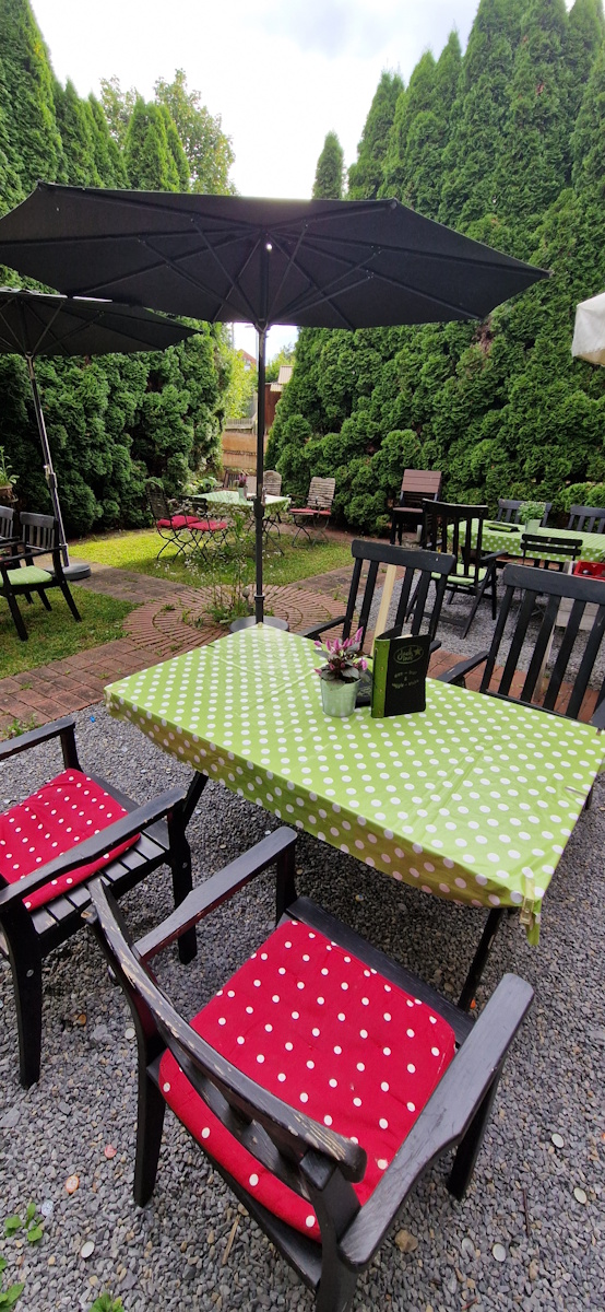 gartenwirtschaft/gartenoase mit tisch und grüne weißgepunktete tischdecke.
                daran stehe 4 stühle mit rot/weißgepunkteten sitzkissen. ein großer sonnenschirm steht am tisch. im hintergrund stehen weiter tische und stühle im selben stil.
                hinter den großen bäumen scheint die sonne durch