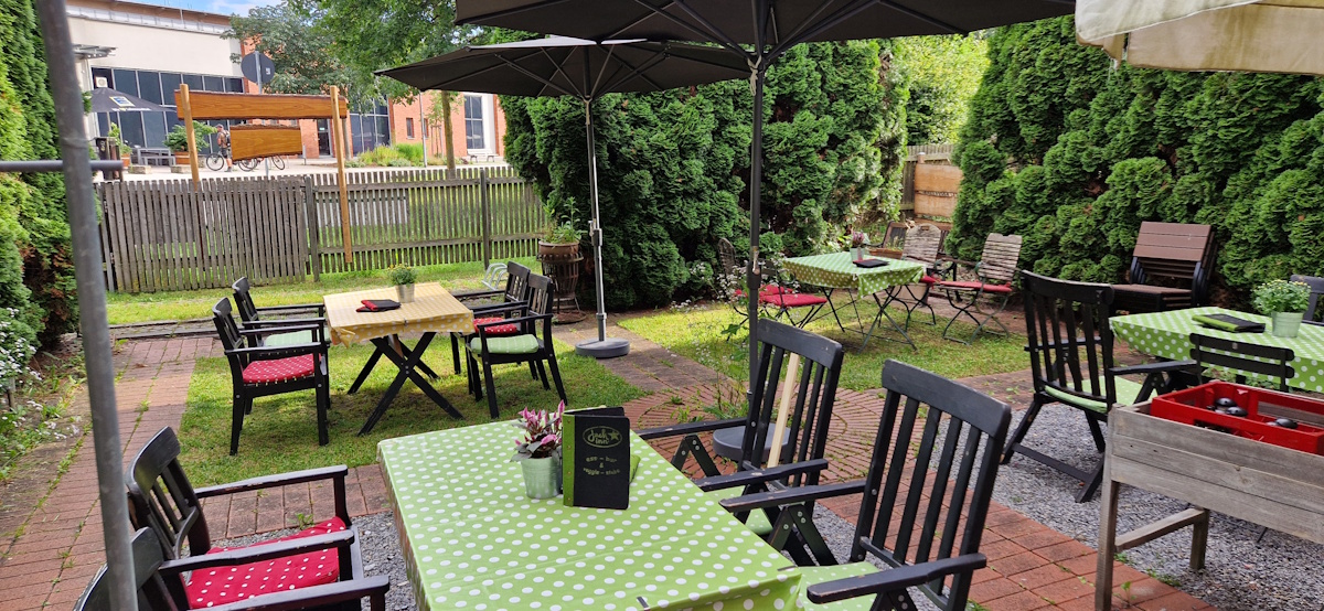 gartenwirtschaft / biergarten mit 4 tischen und stühle an den tischen. 2 große sonnenschirme. im hintergrund sind hohe bäume. es ist ein sommerabend