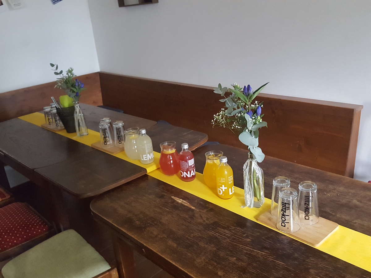 gaststube, alte holztische mit stühlen vor der eckbank. die tische sind aneinander geschoben
                für eine feier eingedeckt. mit gelbem tischläufer auf dem blumen, gläser und limonade stehen,
                2 dekoflaschen mit blumen