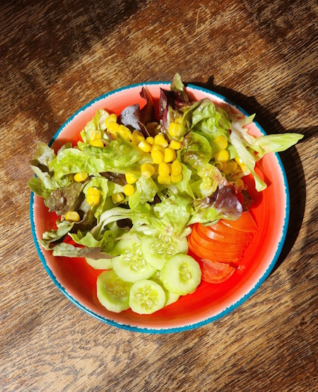 ein kleiner roter teller mit blattsalat, gurken, tomaten und mais auf einem alten holztisch