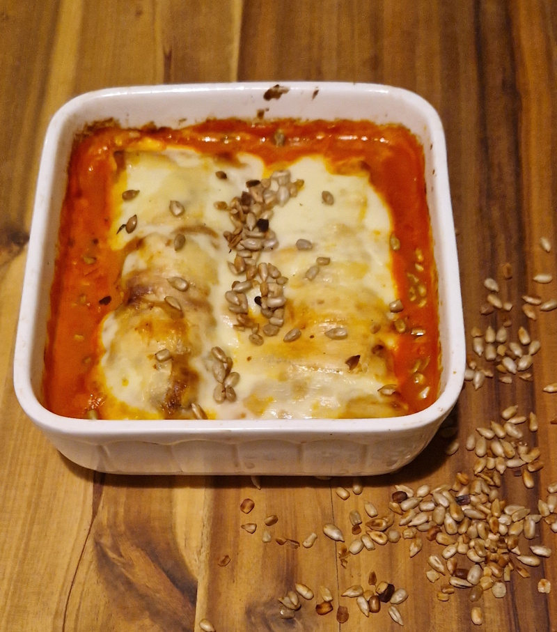 auf einer tischplatte steh eine quadratische weiße auflaufform. dain ist eine tomatensahnesoße zu sehen und
gebackener käse über 2 gefüllten pfannkuchen. es sind geröstete sonnenblumenkerne über dem käse und als deko rechts neben der auflaufform auf dem tisch