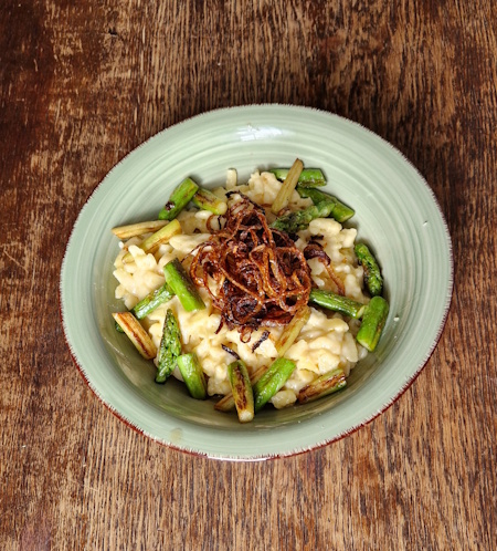 käsespätzle mit frischen röstzwiebeln und gebratene grüne spargel stücke. in einem hellgrünen hohen teller. der teller steh auf einem alten holztisch