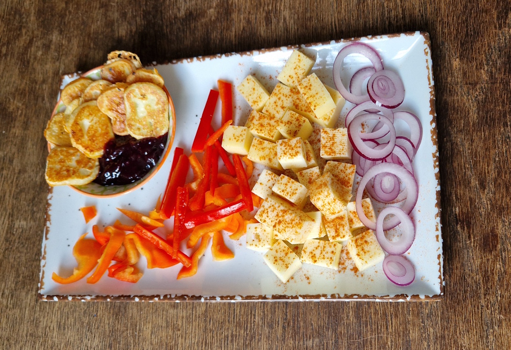 eine weiße rechteckiger teller mit käsewürfel, rote zwiebelringe liegen am rand, rote paprika in streifen ein tellerchen mit fruchtmarmelade und gebratenen camemberttaler,
                die käsewürfel sind mit paprikapulver bestreut. auf einem alten holztisch