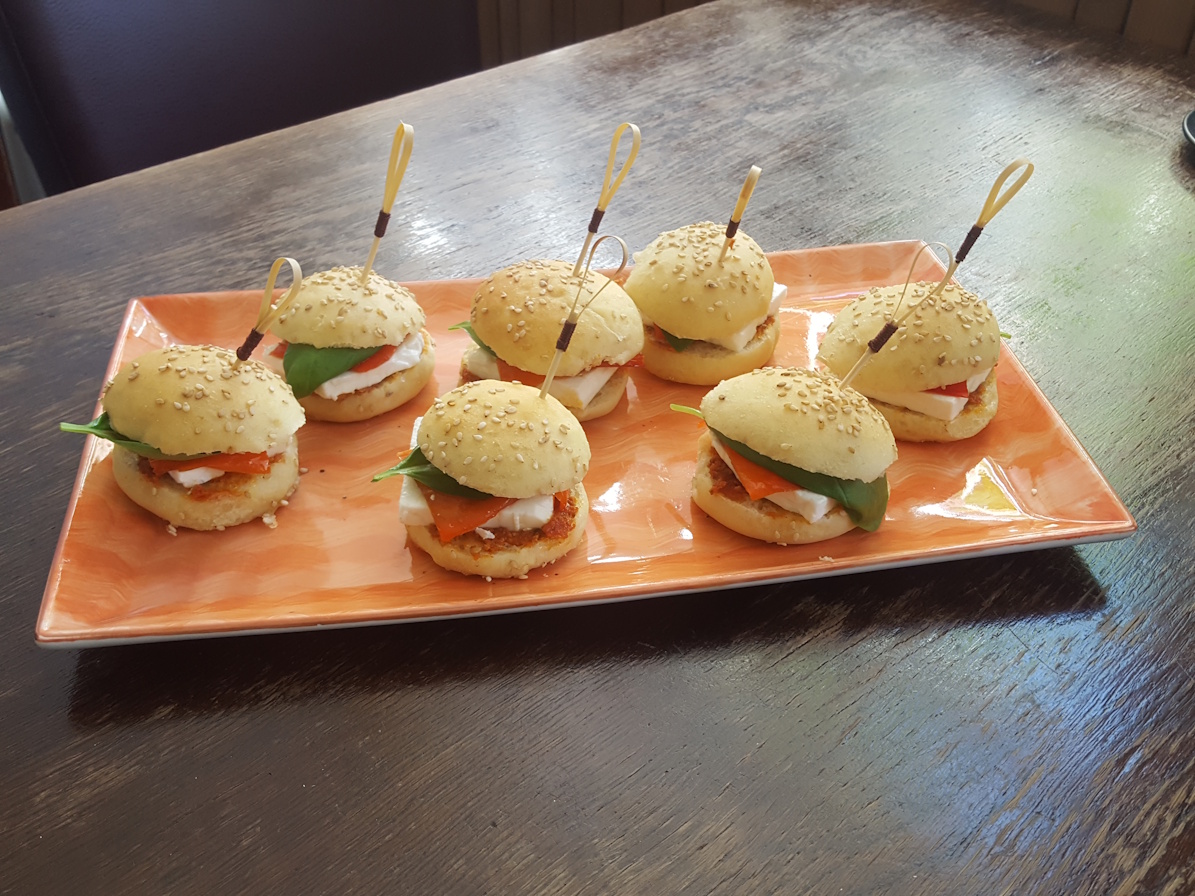 eine kleine platte mit 7 miniburger. belegt mit featkäse, eingelegte paprika und frischem basilikum,
                in den kleinen burgern stecken spießchen zur befestigung. auf einem alten holztisch