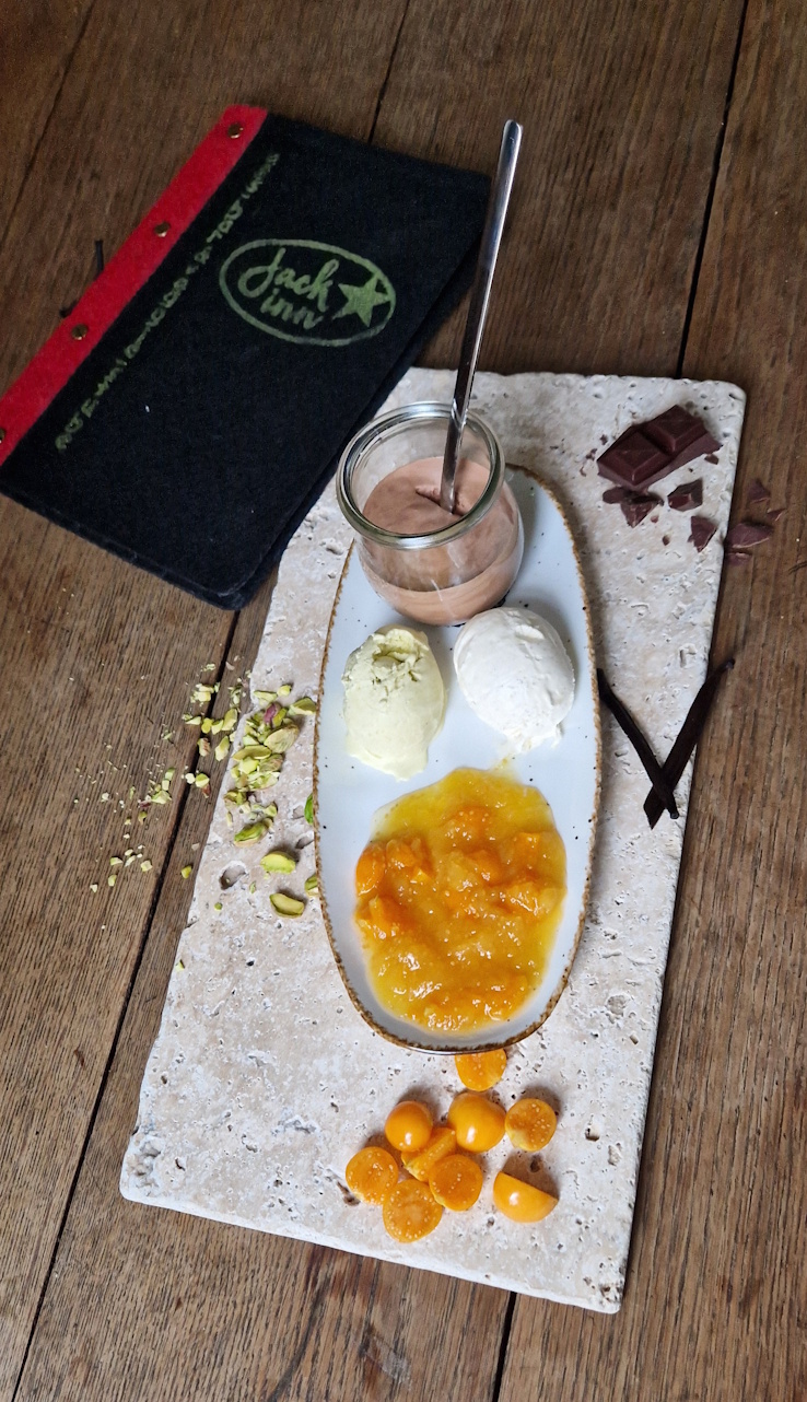 dessert mit mousse au chocolat im glas. je eine kugel hausgemachtes pistazieneis und vanilleeis mit einem frischen physalieskompott auf einer steinfliese. 
                auf einem alten holztisch mit der speisekarte...schwrz mit gelb/grünem aufdruck am oberen rand