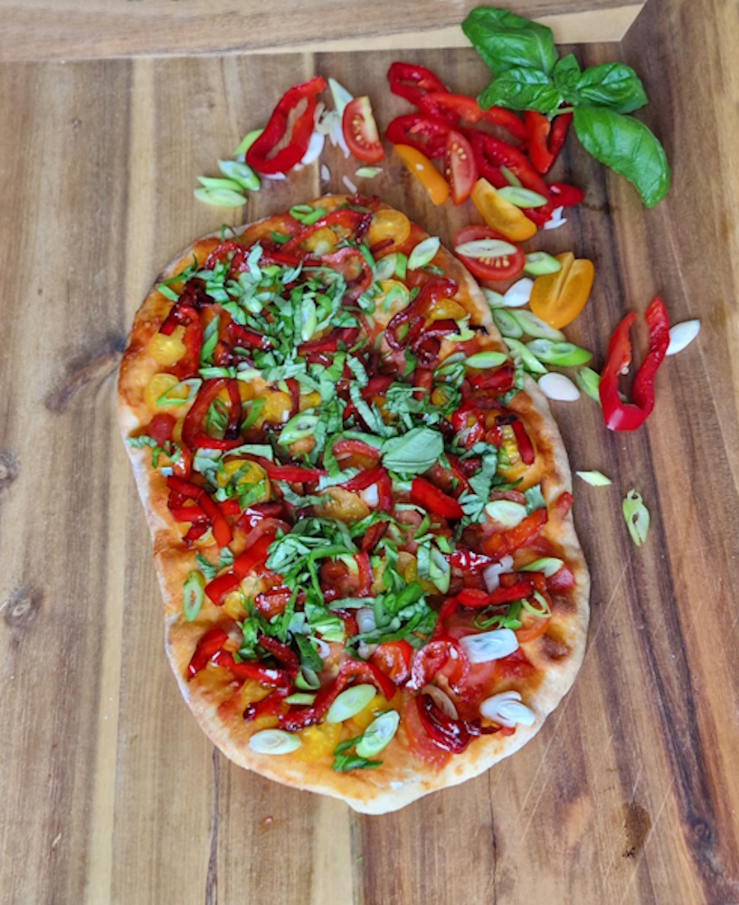 auf einer tischplatte liegt eine belegte pinsa. der belag besteht aus paprikastreifen, lauchzwiebel, halbierte minitomaten
in rot und gelb mit frischem basilikum der in streifen geschnitten wurd. als deko liege die zutaten rechts über der pinsa.