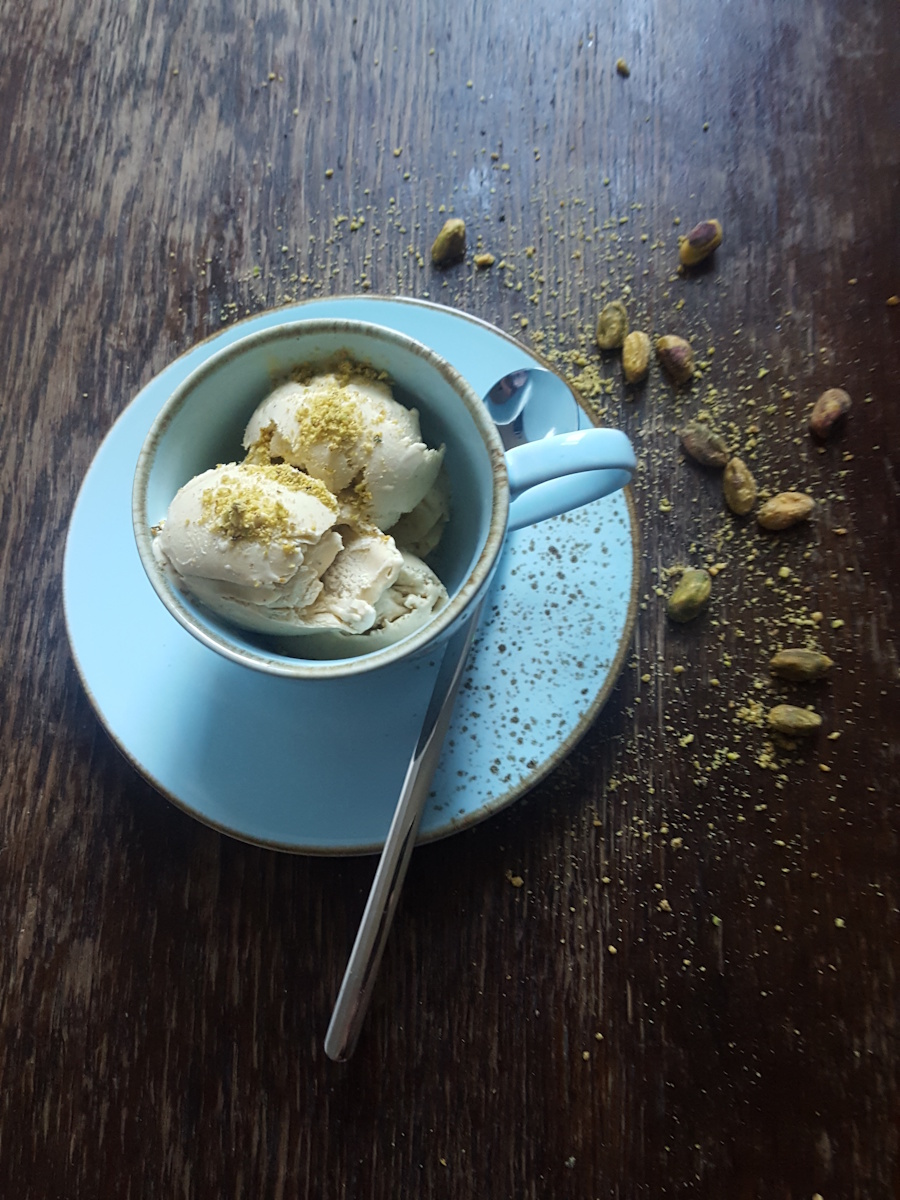 hausgemachtes pistazieneis in einer blaumellierten kaffeetasse mit langem dessertlöffel auf einem alten holztisch,
                dekoriert mit frischen pistazienkerne und zerstoßenen pistazienkerne