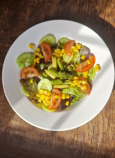 weißer großer salatteller mit blattsalat, tomaten, gurken, mais und gebratene grüner spargel stücke, auf einem alten holztisch