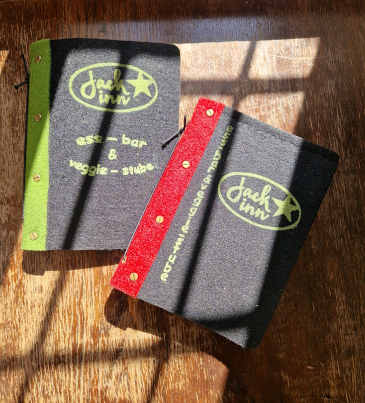 zwei speisekarten aus schwarzenm filz , eine speisekarte mit rotem einband und eine mit grünem einband,
                jeweils mit aufdruck das ovale logo - jack inn- und der aufschrift ess-bar und  veggie-stube. logo und text in gelb/grün. die karten liegen auf einem alten holztisch